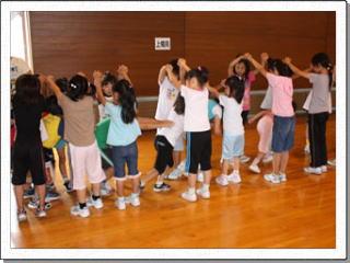 体育館で子供たちが手を頭上でつなぎ、その中を通り抜ける遊びをしている写真