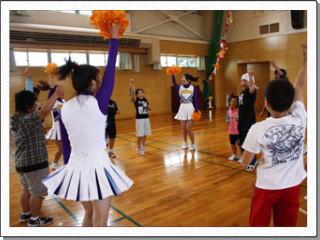 体育館で輪になり手をあげて子供たちと一緒に踊る、チアリーディング部の女性の写真