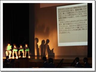 大型スクリーンに文章が映し出され、その左脇で4人並んで緑色のファイルを手に椅子に座る男子生徒のうちの1人が朗読をしている様子の写真
