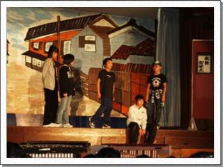 民家と通りが描かれたセットをバックに、5人の児童が演劇を行っている写真