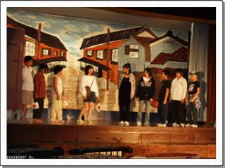 民家と通りが描かれたセットをバックに、日の丸の旗を持った児童など9名が演劇を行っている写真