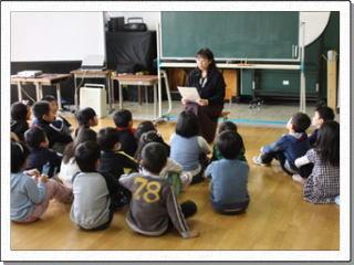床に広がって座る児童たちの前で、資料を手に話をする女性の先生の写真