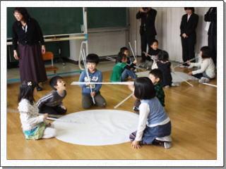 5人の児童がそれぞれに長い箸を持ち床に置かれた白い円の周りに座り、向かいの相手に箸を伸ばしている写真