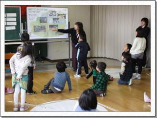 ホワイトボードに貼った資料の説明をしている女性の先生の話を児童たちがそれぞれに立ったり座ったり自由な姿勢で聞いている様子の写真