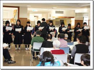 座って聞いている利用者の方たちの前で、横一列に立ち並び楽譜を手に歌っている女子生徒たちの様子の写真