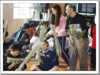 床に座る児童たちに、稲わらの束を手にとって配っている講師と女性の写真