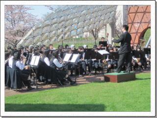 公園に作った簡易ステージでスーツ姿の男性の指揮のもと演奏を披露する緑陵高校吹奏楽部の生徒たちの写真