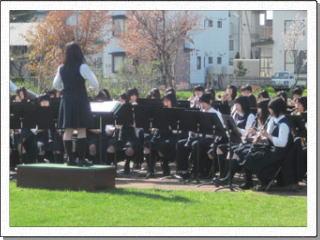 公園に作った簡易ステージでクラリネットやトランペットを構えた部員たちが女子生徒の指揮で演奏を行う様子の写真
