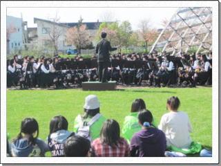公園に作った簡易ステージでの緑陵高校吹奏楽部の演奏を芝生の上に座って鑑賞する人たちの写真