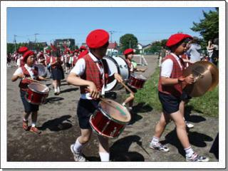 グラウンドを赤いベレー帽と赤いベスト姿の鼓笛隊が列を作って行進している様子の写真