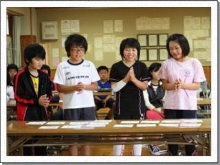 4人の児童たちが教室で紙のカードを並べた机の前に立ち手を合わせている写真