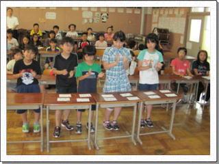 5人の児童が紙のカードを並べた机の前に立ち、手拍子をしている様子の写真