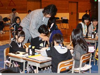 児童4人が机を向かい合わせ座っている席で、1人の児童の手を握り筆の運びを指導する武田双雲さんの写真