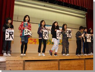 10人程度の児童がステージ上で「沢」と書かれた半紙を両手で持ち横1列に並んで立っている写真