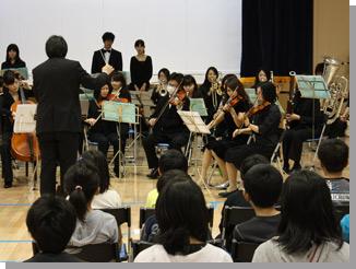 指揮者の男性と楽器を演奏している教育大学岩見沢校音楽コースの学生と演奏に聴き入る生徒達の写真