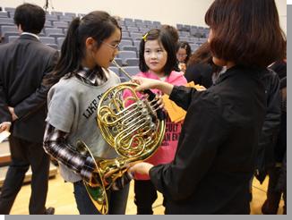 教育大学岩見沢校音楽コースの学生に支えてもらいながらホルンを吹く児童とそれを見る児童の写真