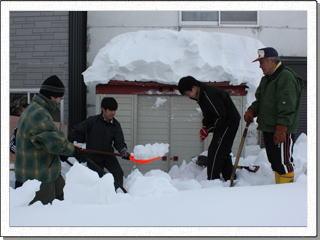 雪が積もった物置と物置前の除雪をする生徒達と男性の写真