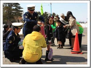 新しいランドセルを背負った男女の児童が警察署員や関係者たちにランドセルカバーをつけてもらっている様子の写真