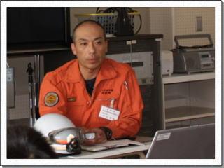 オレンジ色の消防服を着た岩見沢消防署の竹本さんが話をしている様子の写真