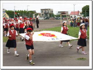 赤いベストを着て赤または白のベレー帽をかぶった4名の児童が学校旗の四隅を持って校庭を歩いている写真
