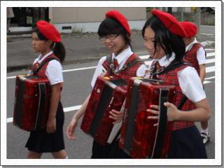 赤いベストを着て赤いベレー帽をかぶった3名の女子児童がアコーディオンを肩にさげて笑顔で道路を歩いている写真