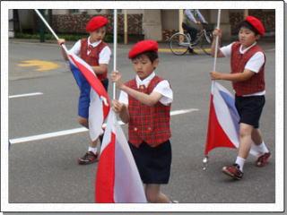 赤いベストを着て赤いベレー帽をかぶった3名の男子児童がフラッグを持って道路を歩いている写真