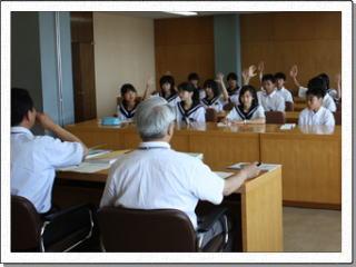 制服姿の複数の生徒たちが、白の半袖ワイシャツ姿の男性2人と机を向かい合わせて座り挙手をしている写真