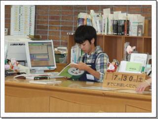 私服に紺色のエプロンを身に着けた男子生徒が書類やパソコンの置かれたカウンターに向かい冊子を手にしている写真