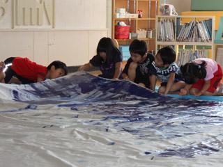 5人の児童が床に跪き色を塗ったカーテンの端を持ち上げ裏側を覗き込んでいる様子の写真