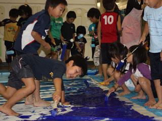 複数の児童が刷毛やローラーを使い青い塗料をカーテンに塗っている写真