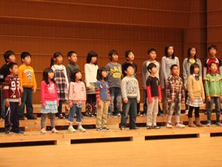 小学校の児童たちがステージ上に2列に並んで立ち、合唱をしている様子の写真