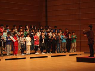小学校の児童たちがステージ上に3列に並んで立ち、ピアニカやリコーダーで合奏している様子の写真
