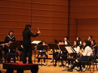 中学校の生徒たちがステージ上でクラリネットやフルートで吹奏楽を披露している様子の写真