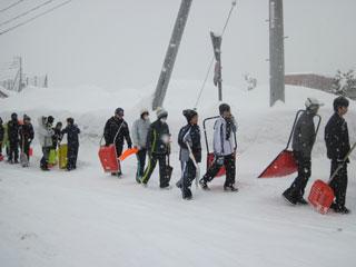 スコップを持った生徒たちが雪の積もった道路の脇を一列になって歩いていく様子の写真