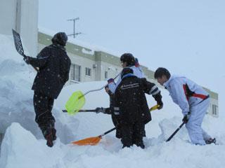 胸の高さくらいまで積もった雪を腰を入れてスコップを使って掻き出している4人の生徒たちの写真