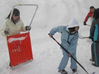 水色の服を着た女の子がスコップを使って雪かきをしている横で、体の半分くらいの大きな赤いスコップを立てて持っている女の子の写真