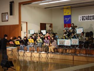 体育館のステージ上にパイプ椅子に座って並び楽器を演奏している緑中学校吹奏楽部の生徒たちの写真