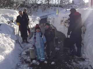 3人の男性が雪をかき分けて作った道を、小さな男の子と女の子が並んで歩いている写真