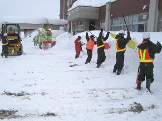 カラフルな作業着を着た5人の男性が、スコップを使って校舎脇に雪を身長くらいの高さまで積み上げている様子の写真