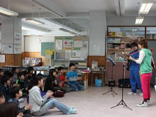 教室で翔太さんと先生がギターを持って立ち、床に座った子どもたちが拍手をしている様子の写真