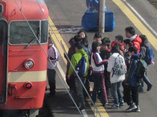 15名程度の児童たちがホームでかたまり、停車した電車に乗り込もうとしている様子の写真