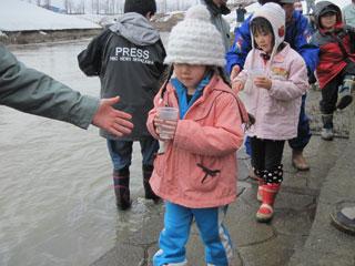 ピンクのコートに、灰色のニット帽を被った女の子が大事そうに稚魚の入ったコップを抱えている写真