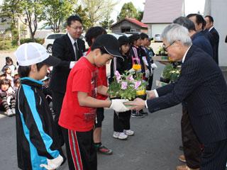 黒い帽子をかぶり、赤いシャツを着た男の子がスーツの男性から花を受け取っている写真