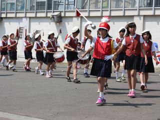 先頭でバトンを振る女子児童に続き複数の児童が楽器を構えながらパレードに出発する様子の写真