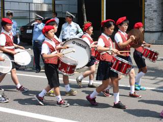 赤のベレー帽をかぶった7名の児童たちが、それぞれに大太鼓と小太鼓、シンバルをたたきながら行進している写真