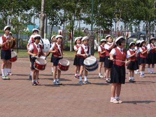 白いベレー帽をかぶった児童たちがバトンを振る女子児童を中心に扇形に並び楽器を演奏している写真
