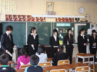 6名の大学生が机に座る児童たちに向かい楽器を手に黒板の前に立っている写真
