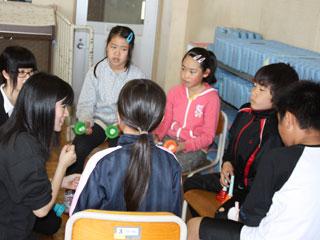 2人の女子大生と5人の児童がハンドベルを手に持ち輪になって話し合う様子の写真