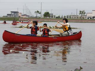 赤いカヌーに生徒二人と一人の女声が乗り込み、オールを振って漕いでいる様子の写真