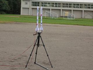 学校のグラウンドで三脚に固定された手作りロケットの写真